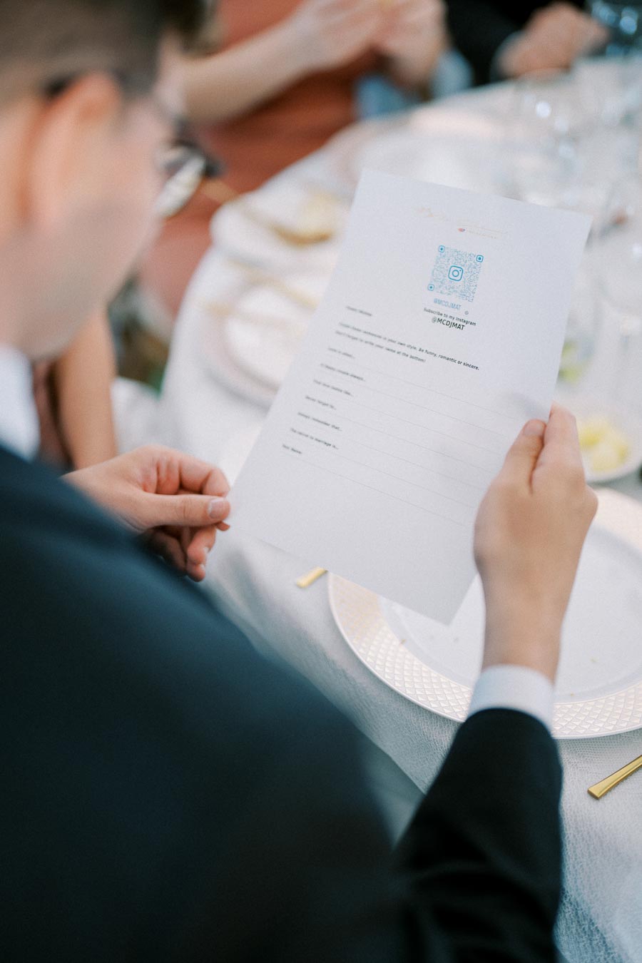 A person in formal attire reading a menu or document at an elegantly set dining table with plates and glassware in the background.
