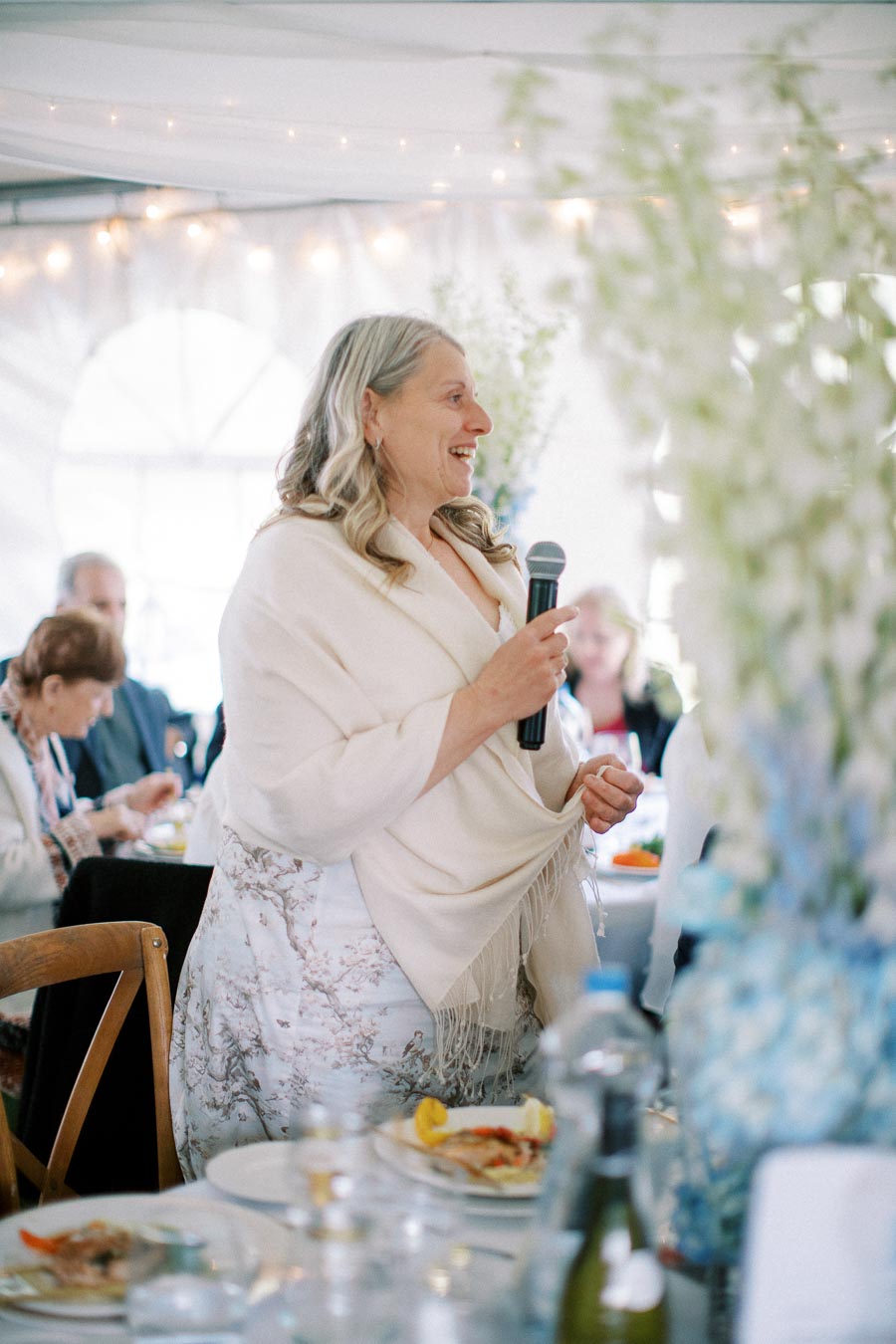 Elderly woman giving a speech at an elegant indoor event, holding a microphone and smiling, surrounded by seated guests and decorative lights.