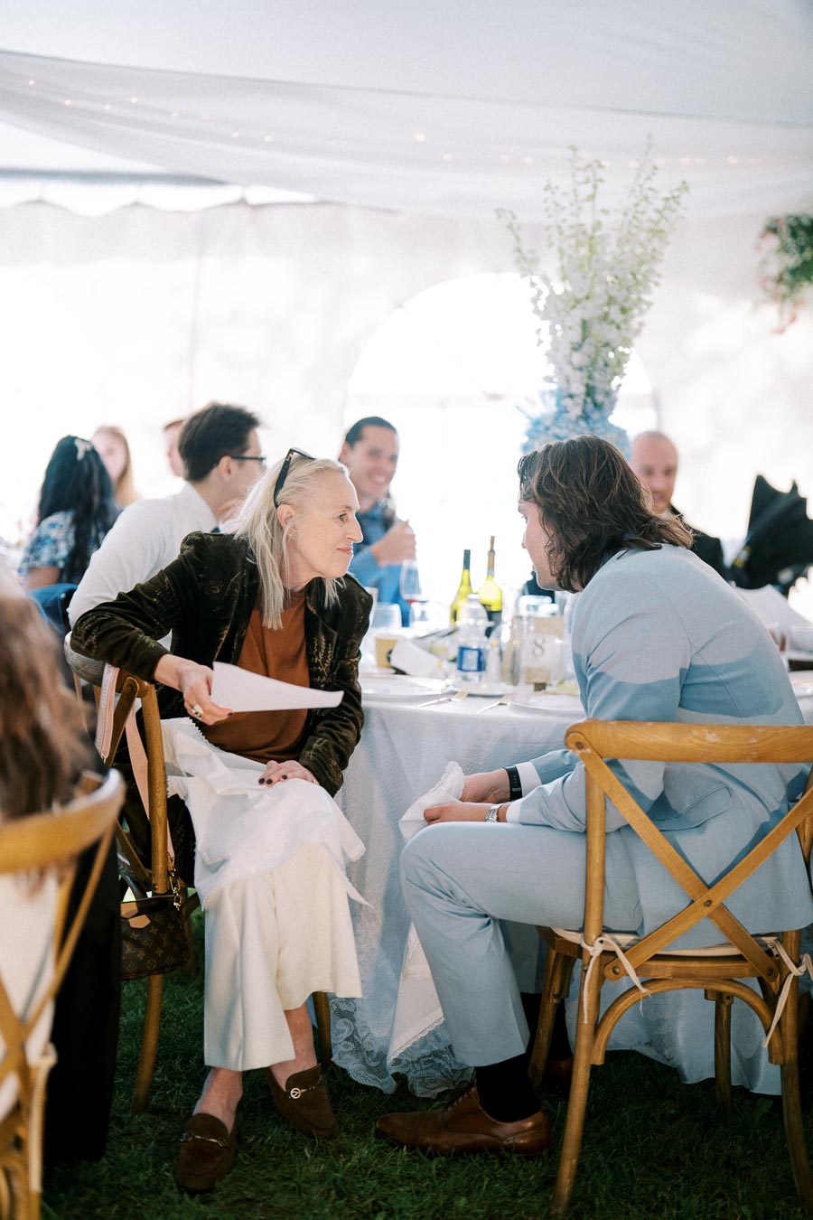 Guests seated at a beautifully decorated outdoor event, engaged in conversation. Tables are adorned with white tablecloths, wine bottles, and floral arrangements under a white canopy.