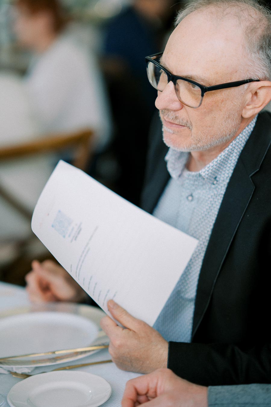 A person in glasses and a suit holding a document while sitting at a table, engaged in a conversation or meeting setting.