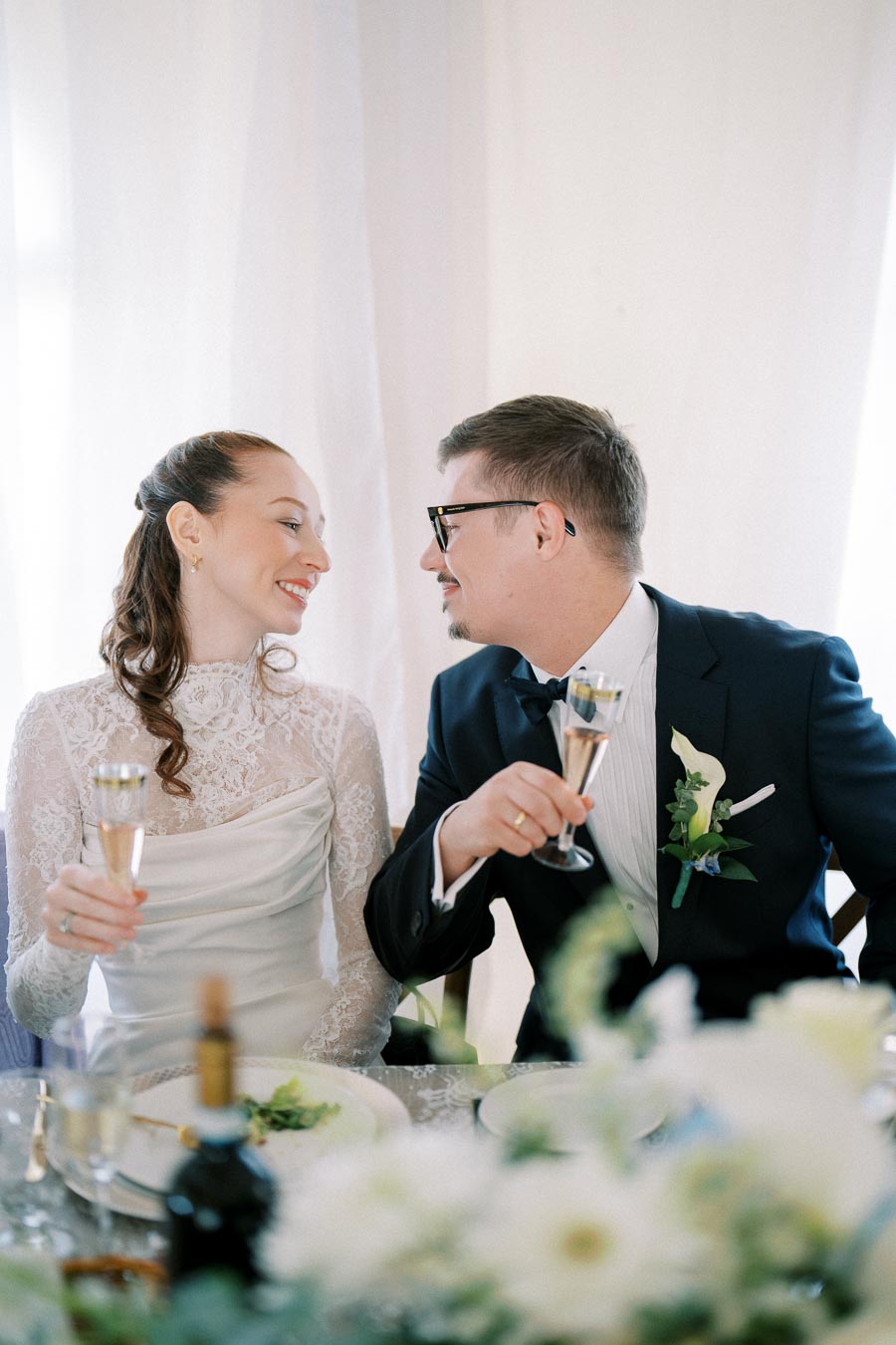 Bride and groom clinking champagne glasses, smiling at each other during wedding reception with elegant floral decor in foreground.