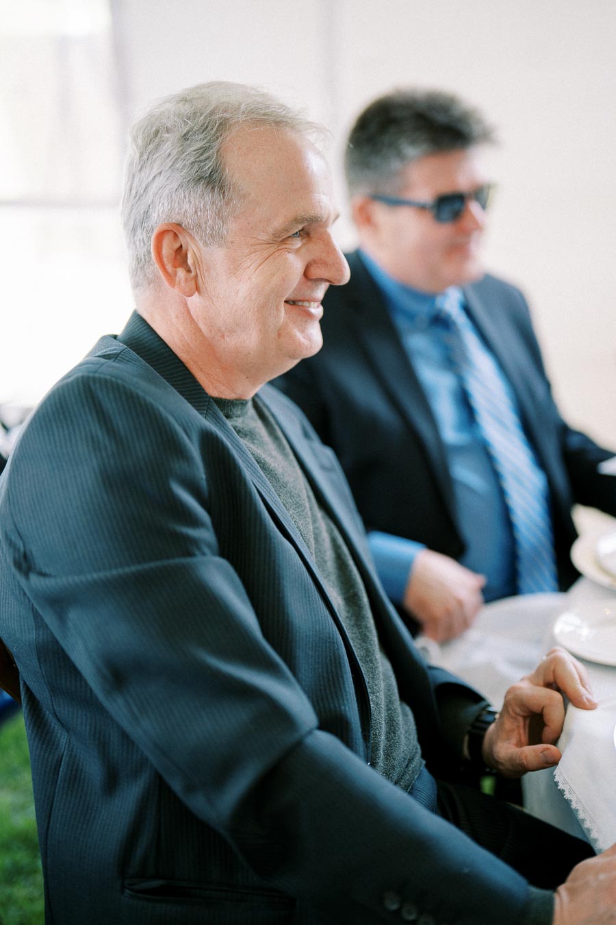 Two men in suits sitting at a dining table, one smiling and leaning forward, suggesting a professional gathering or business event.