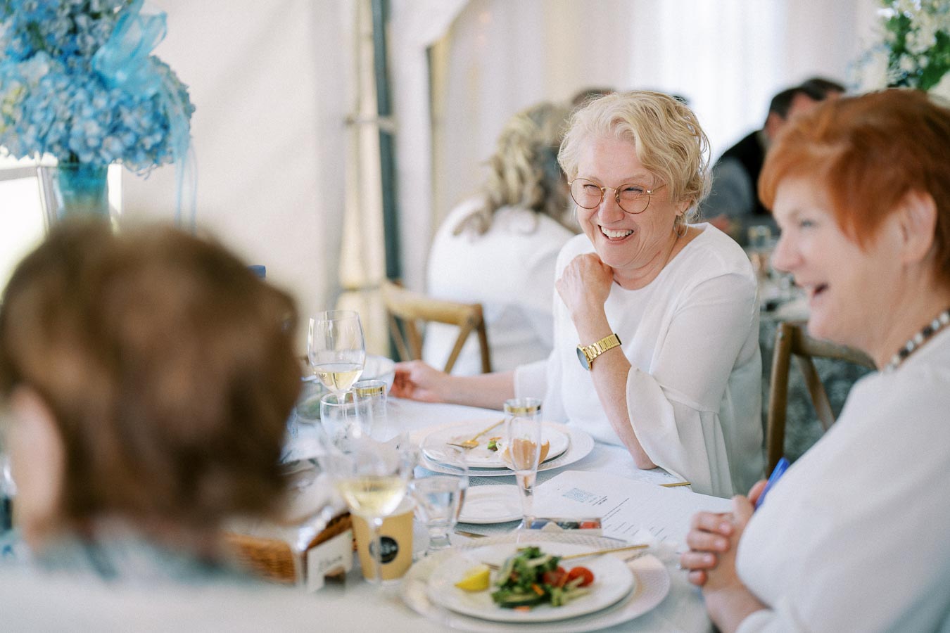 Elderly women enjoying a meal and conversation at a beautifully decorated dining table with blue flowers and wine glasses.