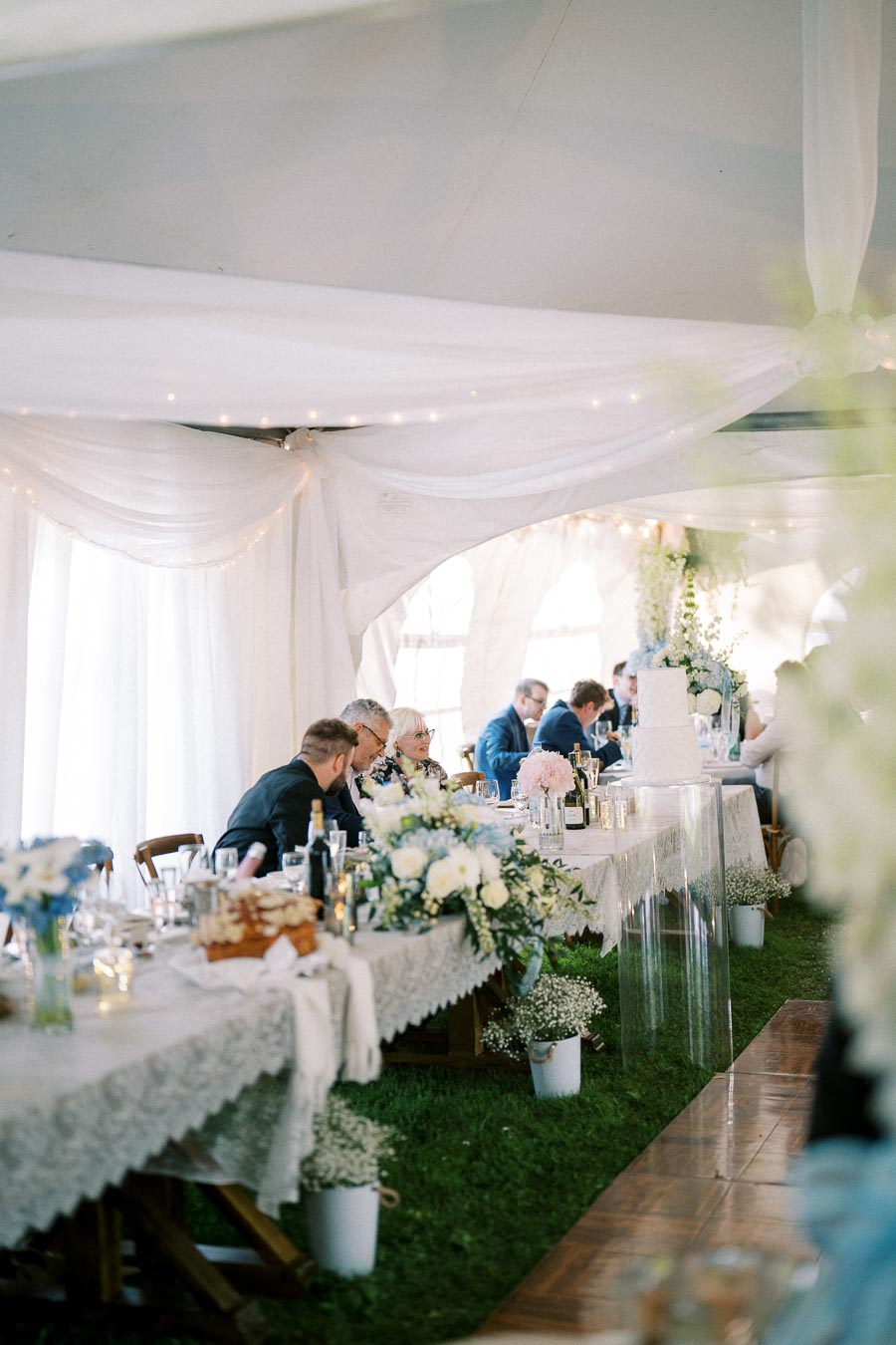Elegant wedding reception in a beautifully decorated marquee with guests seated at tables adorned with floral centerpieces and white drapery.