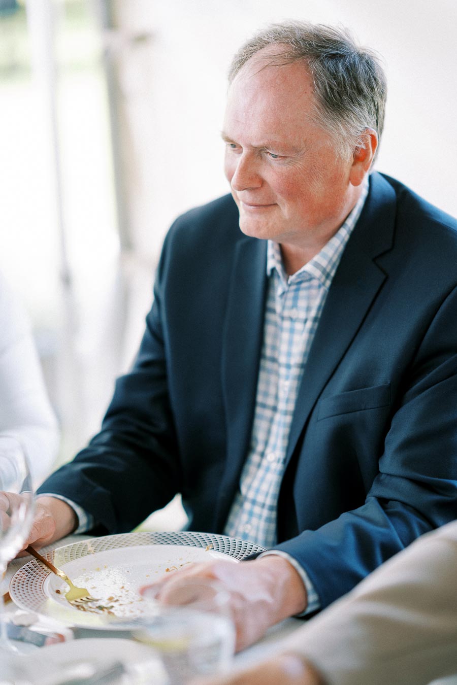 A man in a navy blazer and checkered shirt enjoying a meal at a formal gathering.