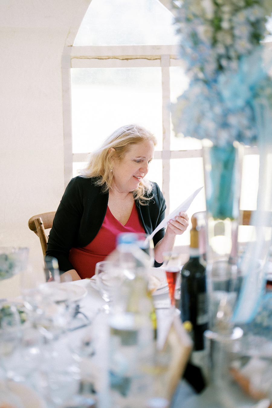 A woman in a black jacket and red dress sits at a dining table, reading a piece of paper. The table is elegantly set with glassware, wine bottles, and blue floral arrangements in a bright, airy setting.