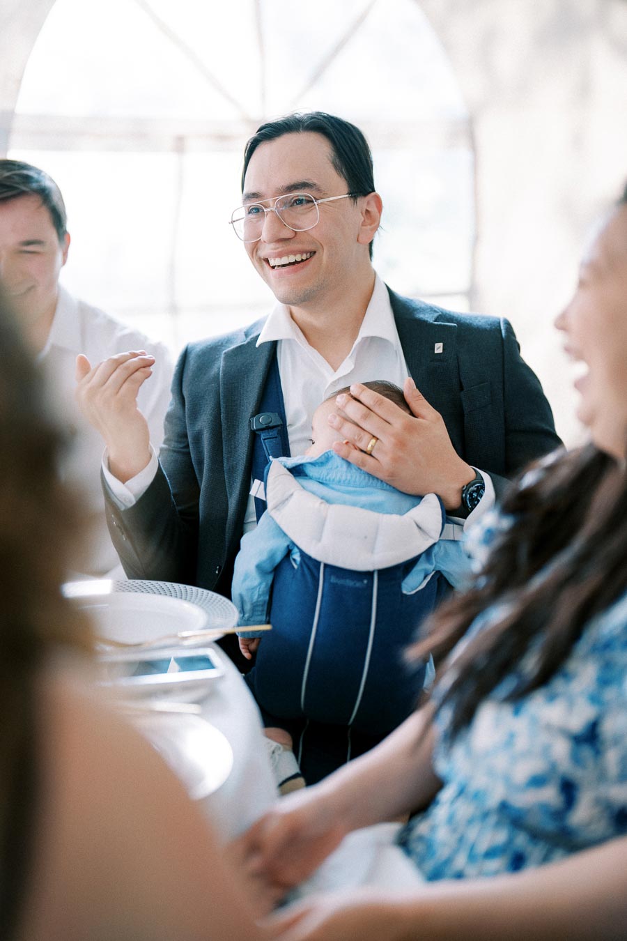 Man in a suit smiling and holding a baby in a carrier, engaging in a joyful conversation at a gathering or event.