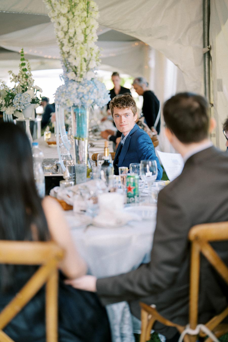 Elegantly dressed man in a suit at a decorated outdoor event, seated at a table with floral centerpieces and drinks, under a large tent setting.