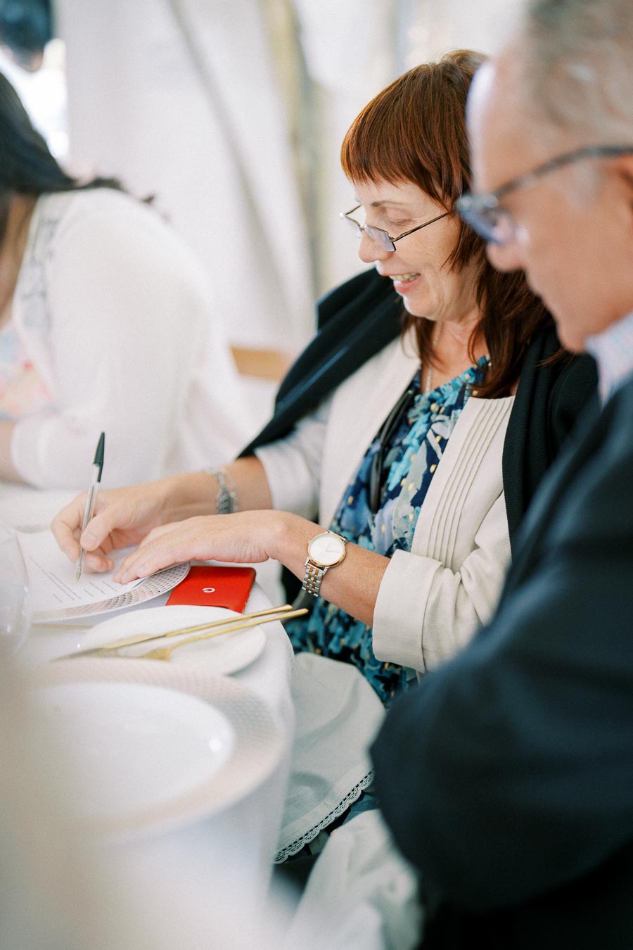 Woman smiling while writing on a document at a formal event, seated at a table with tableware and gold utensils.