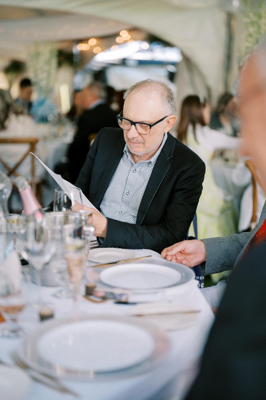 An older man wearing glasses reads a menu at a formal dining table set for an event, surrounded by elegantly dressed guests in an ambient setting with soft lighting.