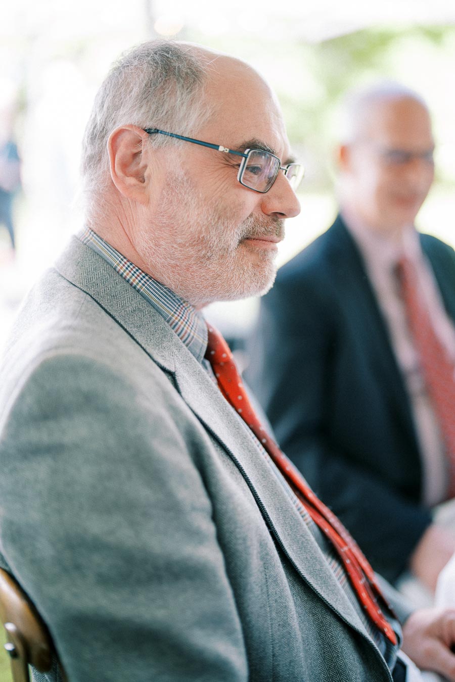 A distinguished gentleman with a beard and glasses wearing a grey suit and red tie, sitting at an outdoor event with blurred greenery in the background.
