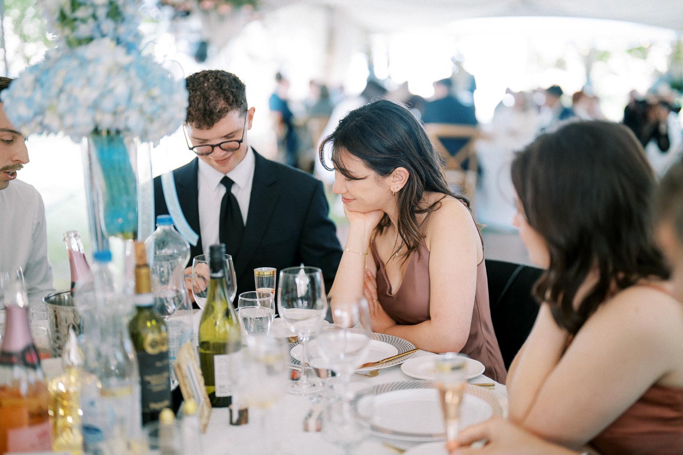 Guests enjoy a formal occasion, dressed elegantly, sitting around a table adorned with wine bottles, glasses, and floral centerpieces at a daytime event.