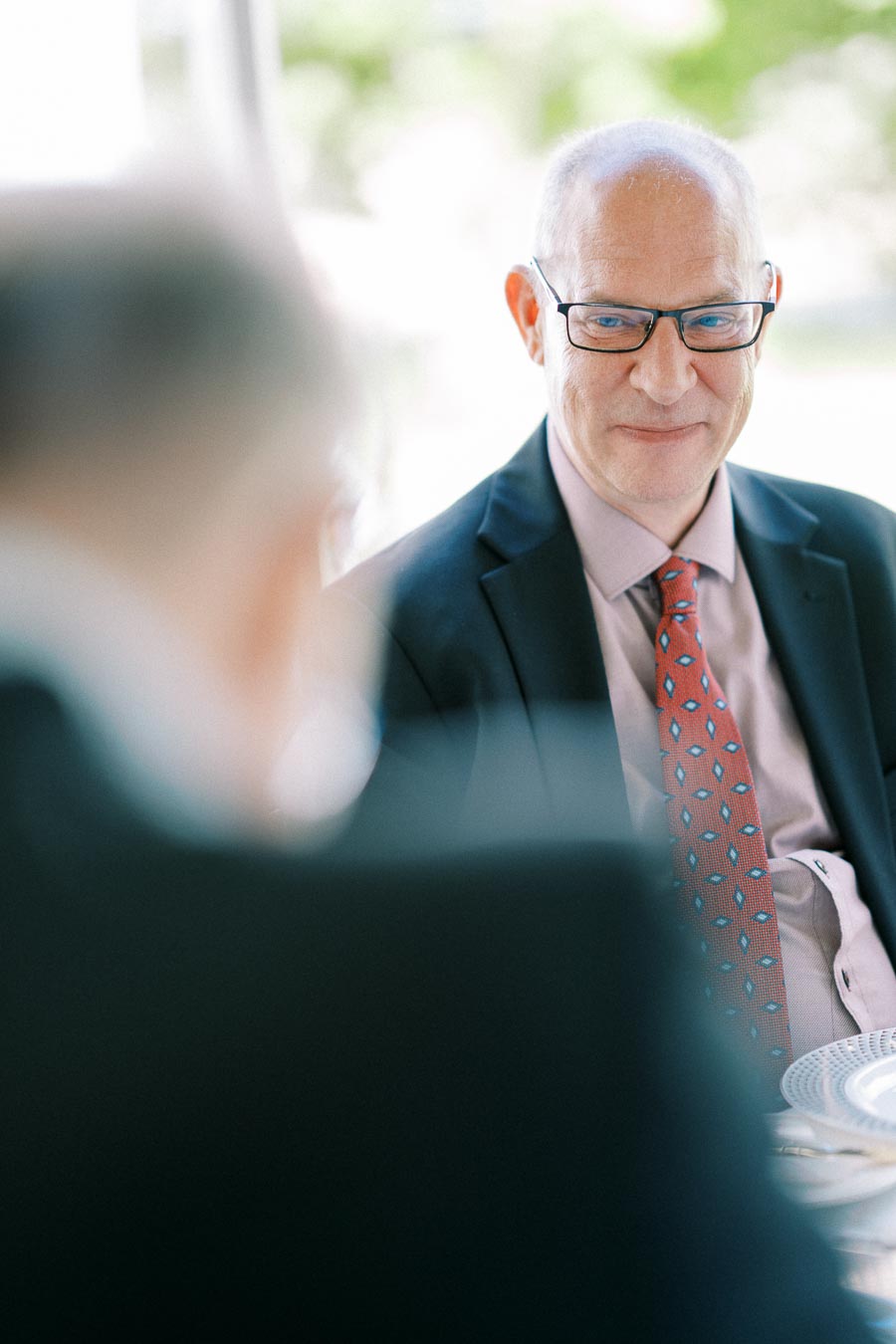 Senior man in glasses wearing a suit and patterned tie, sitting at an outdoor event with blurred figures in the foreground.