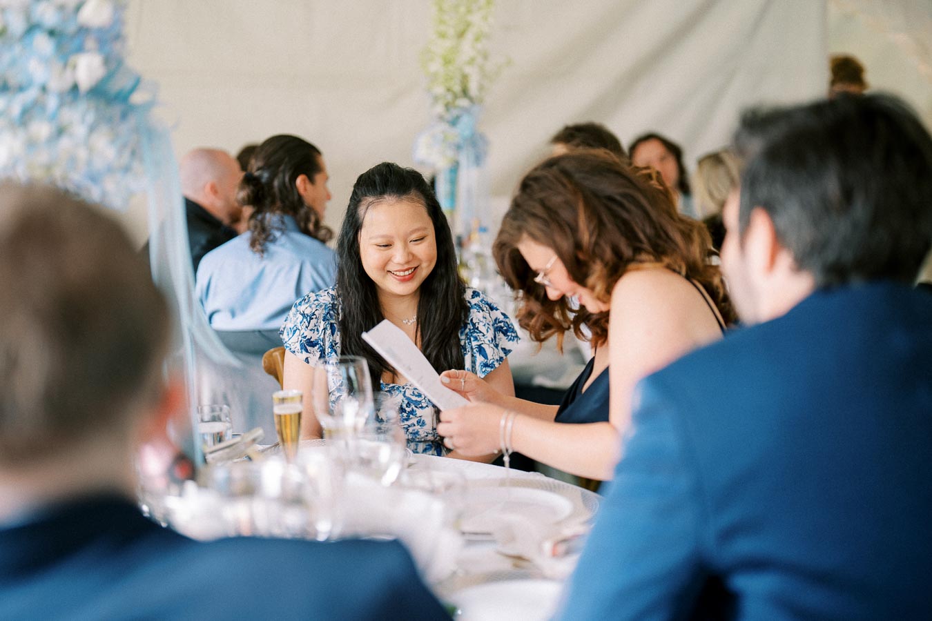 Group of people enjoying a wedding reception, with two women at the center reading a card at a beautifully set table.