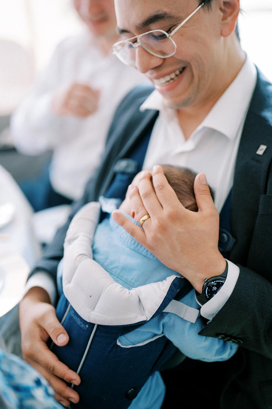 A smiling man holding a sleeping baby in a blue carrier, wearing glasses and a gold ring, conveying happiness and family bonding.