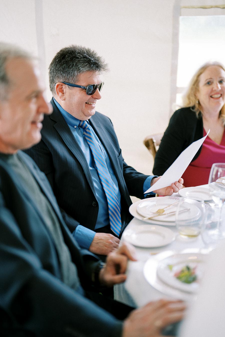 Guests smiling and enjoying a formal event while sitting at a table set with plates and glasses, with one person wearing sunglasses and reading a document.