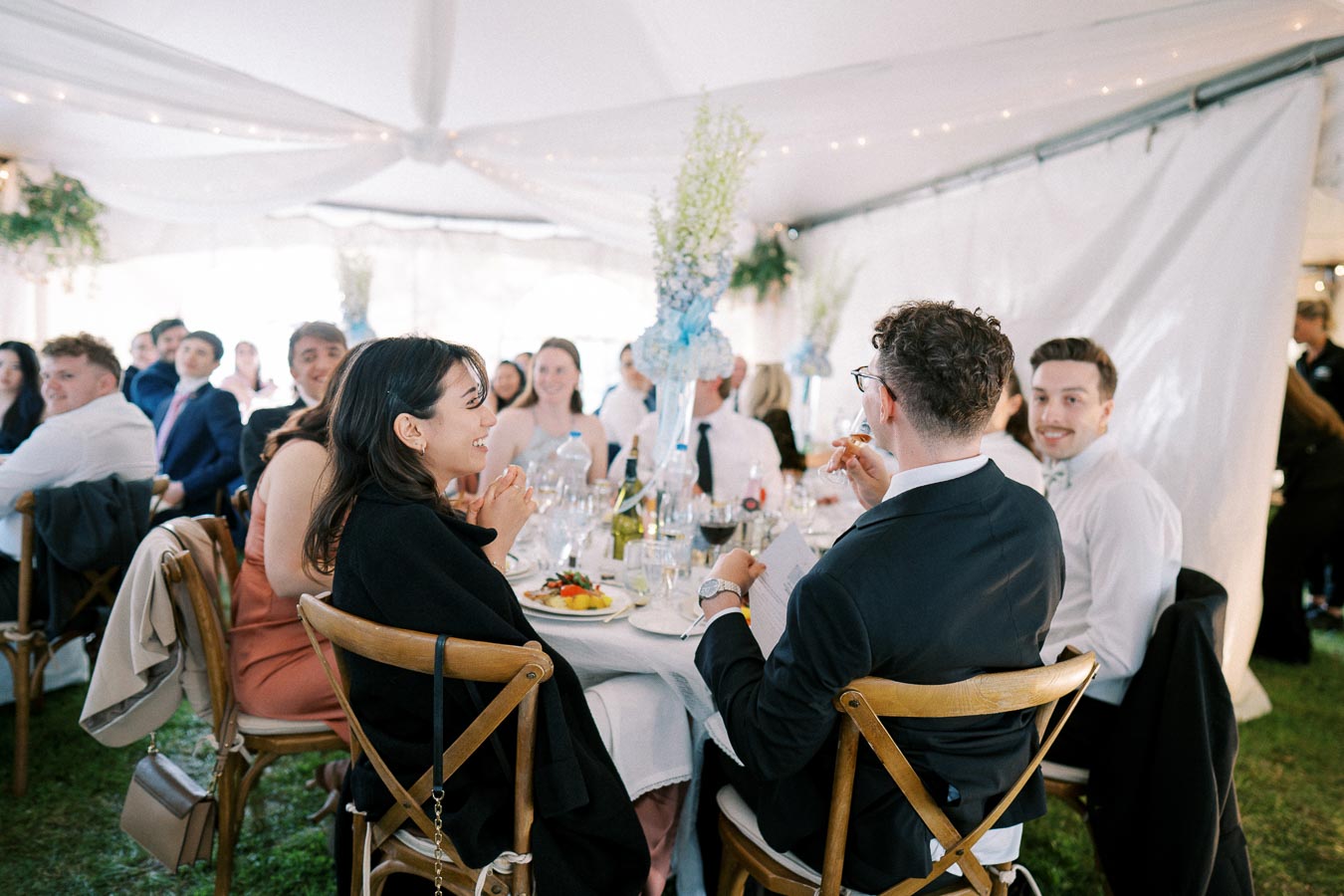 Group of well-dressed people enjoying a formal event at a decorated indoor venue, seated around tables with elegant place settings and floral centerpieces.