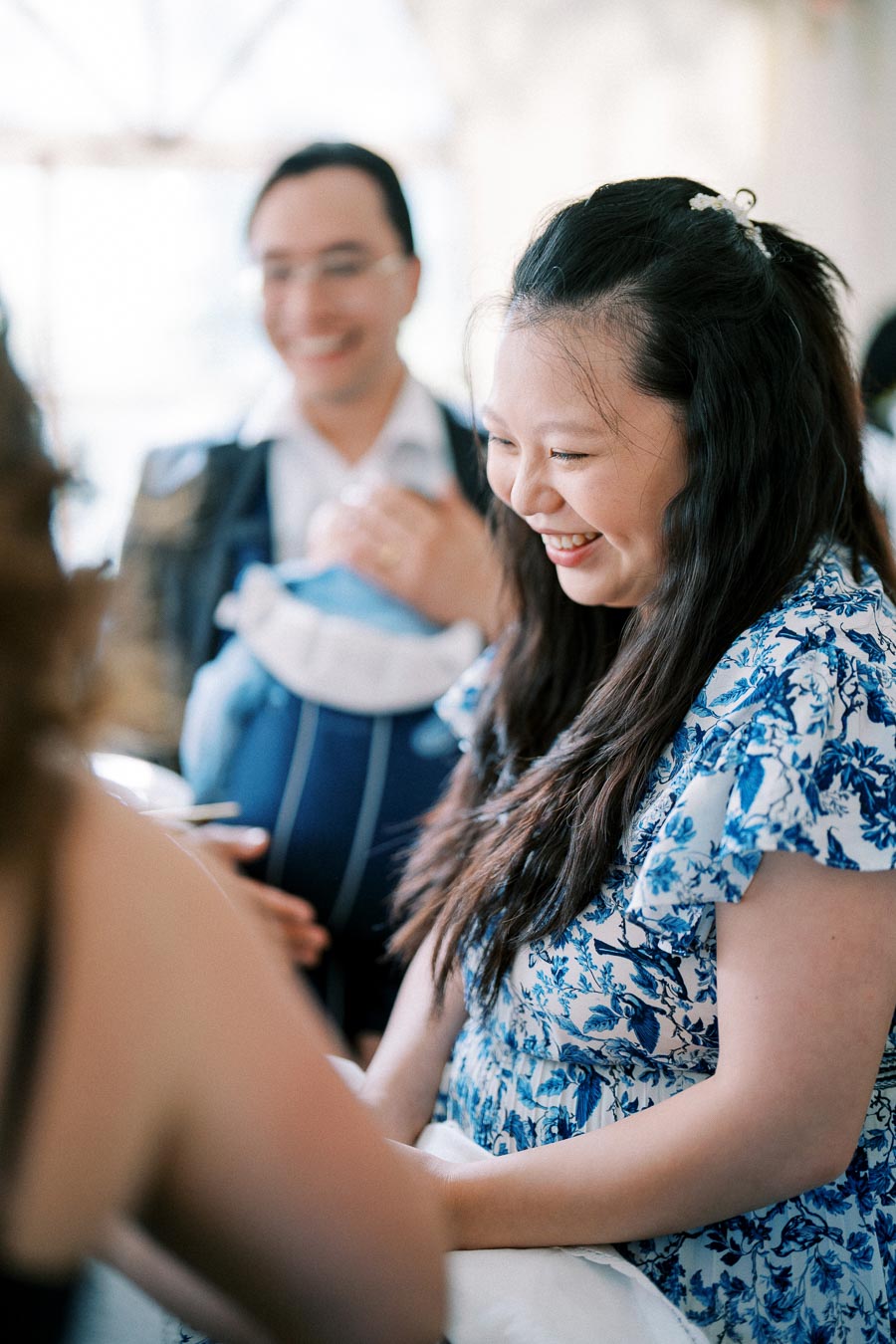 Smiling woman at a social gathering, wearing a blue floral dress, interacting with others in a bright, airy setting