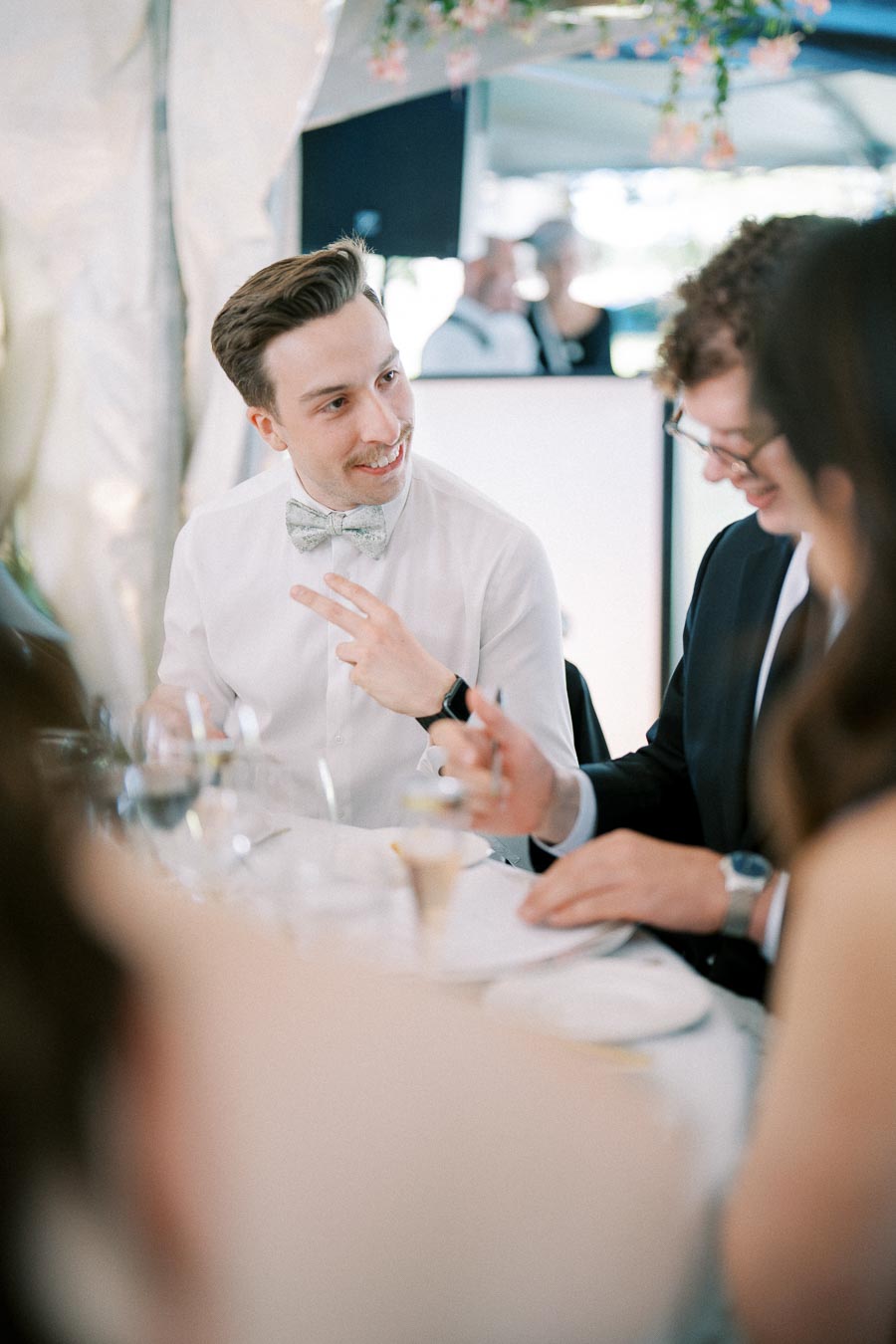 A man in a white shirt and bow tie smiling and chatting with friends at an elegant dinner event.