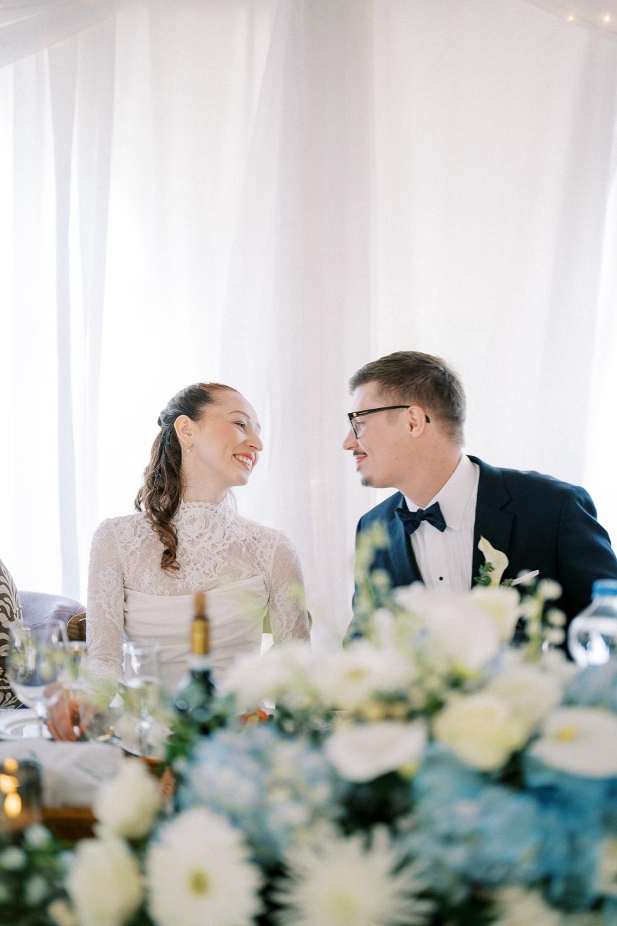 Bride and groom smiling at each other during their wedding reception, surrounded by elegant floral arrangements in soft white and blue hues.