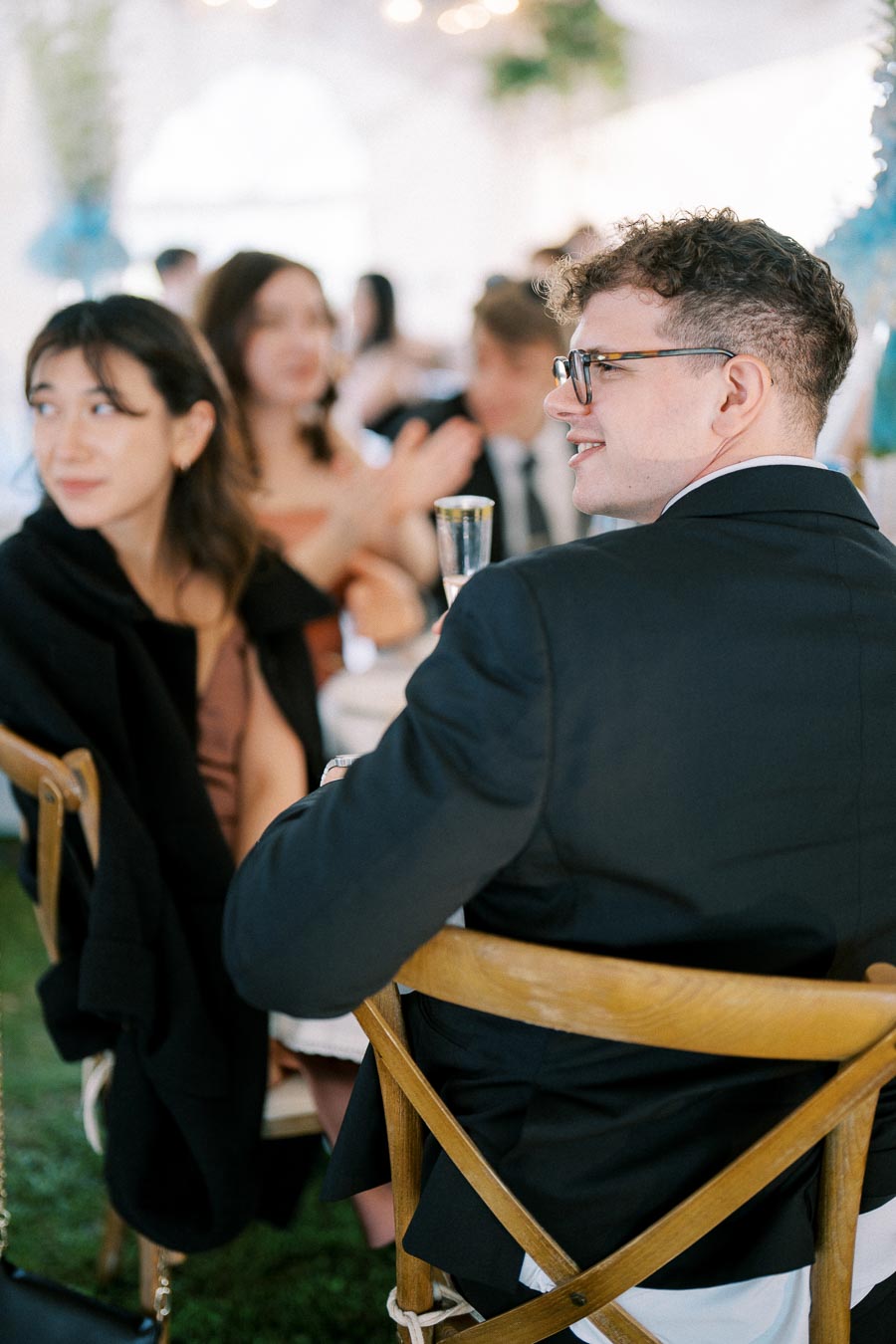 Guests seated at a formal event, engaging in conversation and dressed in elegant attire, with a festive atmosphere highlighted by blurred lights in the background.
