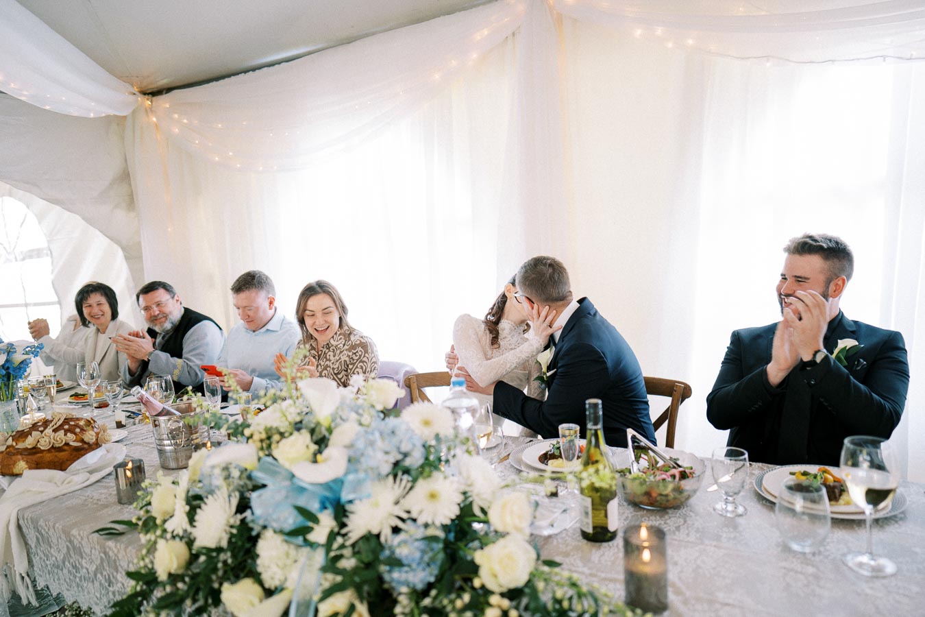 A wedding reception under a softly lit tent with a long table adorned with white and blue floral arrangements. Guests smile and clap as the bride and groom share a kiss, creating a joyful and celebratory atmosphere.