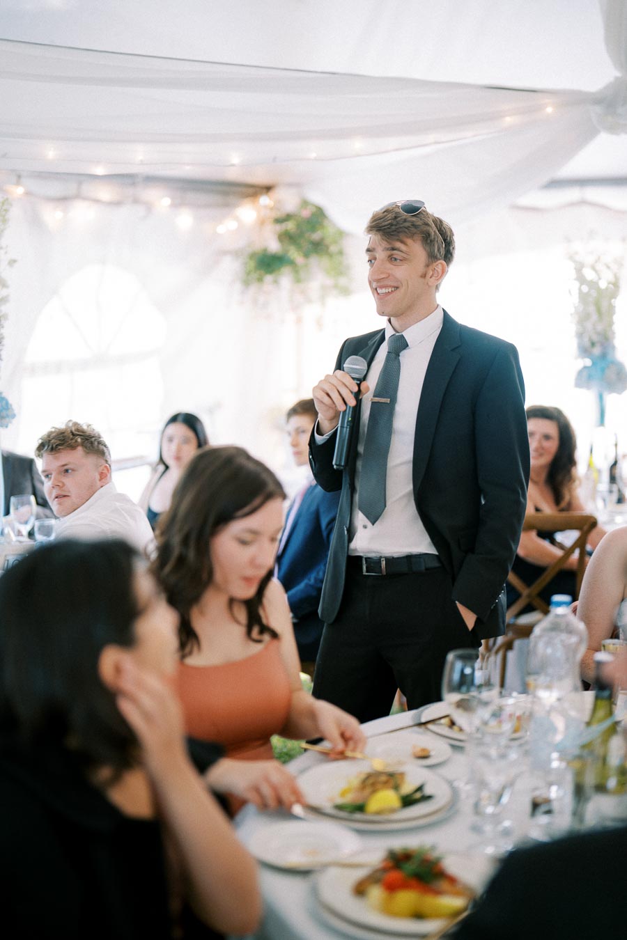 A man in a suit giving a speech at a wedding reception, holding a microphone and smiling, surrounded by seated guests at a dining table with plates of food and drink, under a draped tent with decorative lights.