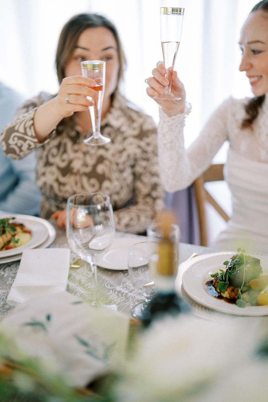 Two people raising champagne glasses in a celebratory toast at an elegantly set table with plated gourmet food and wine glasses, during a festive celebration or wedding reception.
