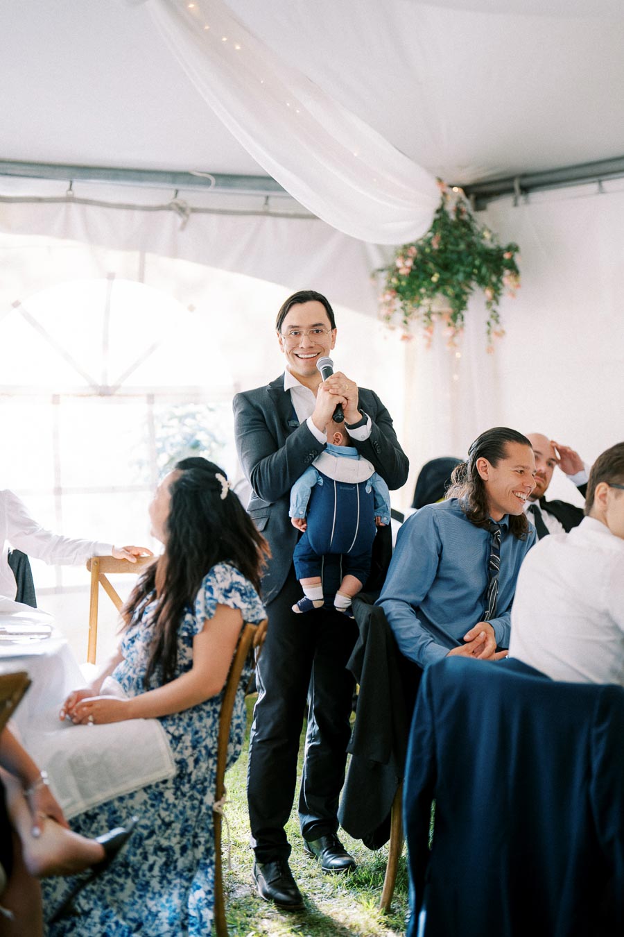 Man in a suit smiling while holding a microphone and a baby at a formal event with seated guests and decorative drapery in the background.
