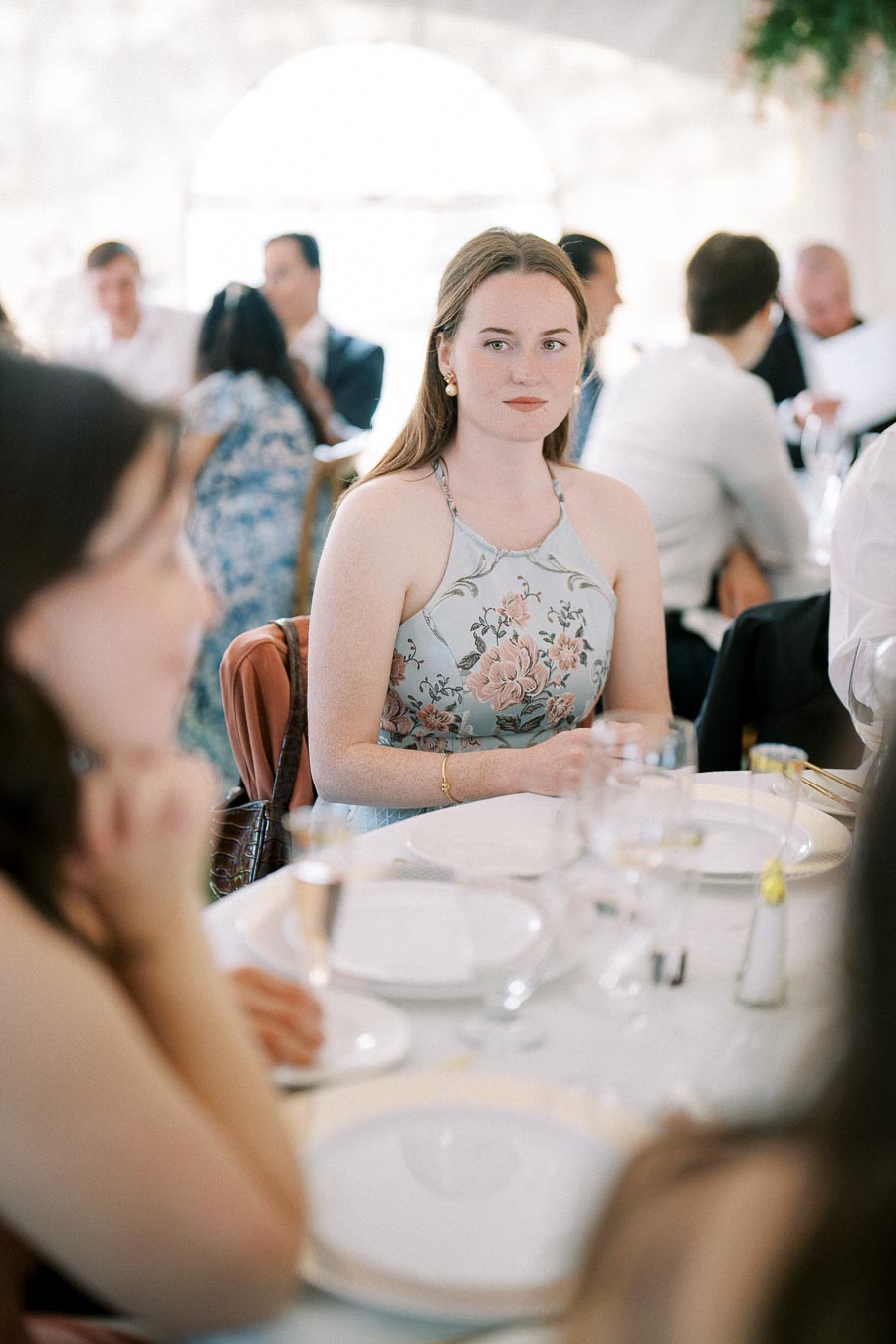 Woman in floral dress seated at a formal dining event with other attendees in a softly lit setting.