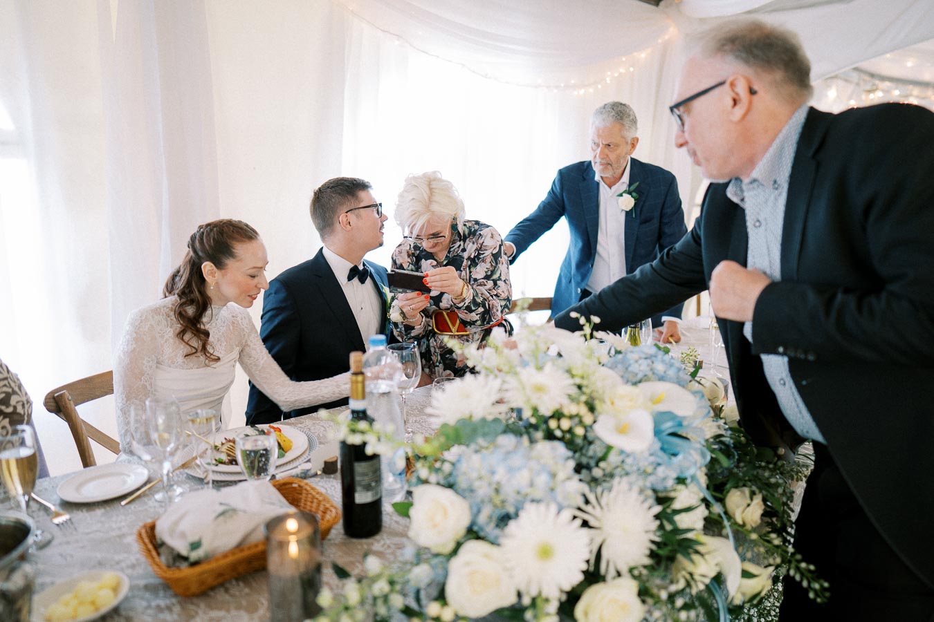 Wedding reception scene with a bride in a white dress and a groom in a suit seated at a table, surrounded by guests. One guest is showing something on a phone, while others engage in conversation. The table is elegantly set with floral arrangements and dinnerware.