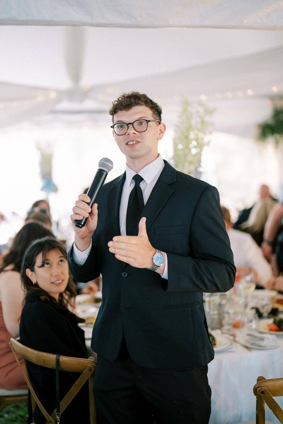 A man in a suit holding a microphone, speaking at a formal event. A woman sits attentively at a table with food and drinks in the background. The setting appears to be a wedding or banquet under a tent.
