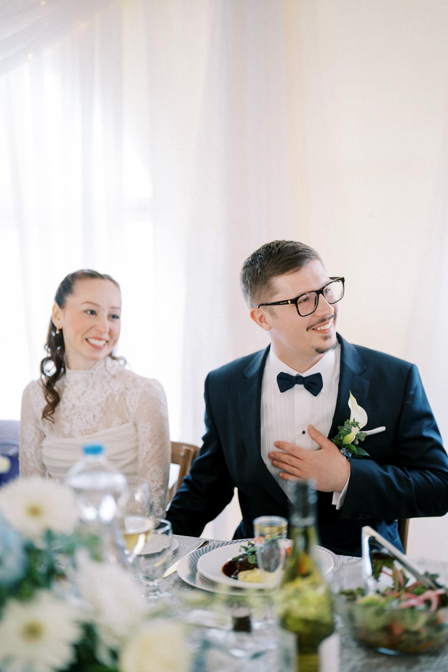 Smiling bride and groom sitting at a beautifully set wedding reception table with elegant floral arrangements