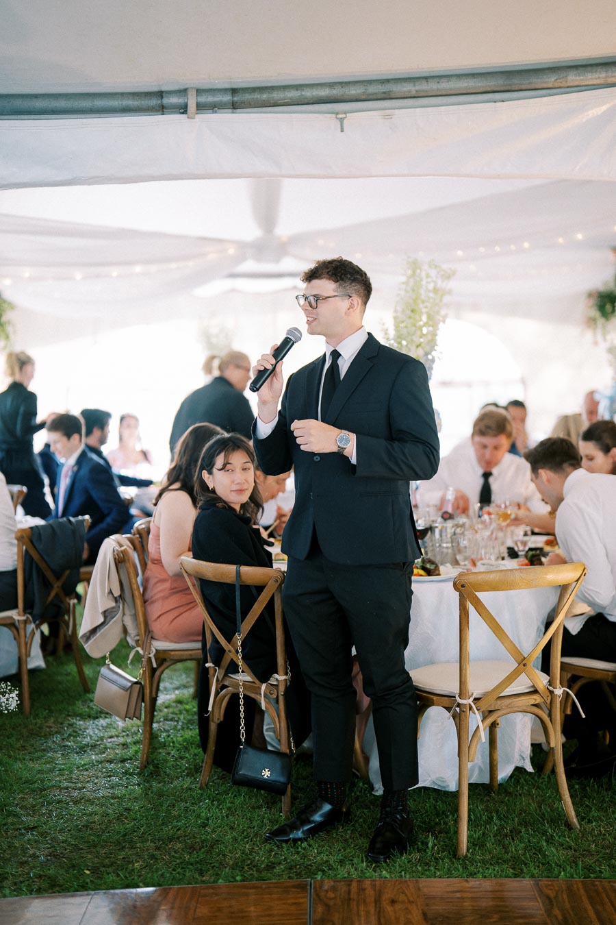 A well-dressed person giving a speech with a microphone at a formal event under a white tent. Guests are seated at round tables with white tablecloths, enjoying a meal and listening attentively.