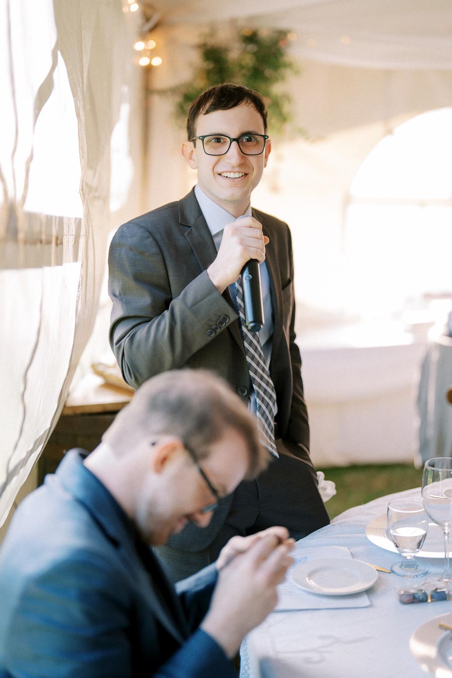 A man in a suit with glasses holds a microphone, smiling at a formal event. Another person is sitting at a table in the foreground, out of focus, suggesting a joyful occasion like a wedding or celebration.