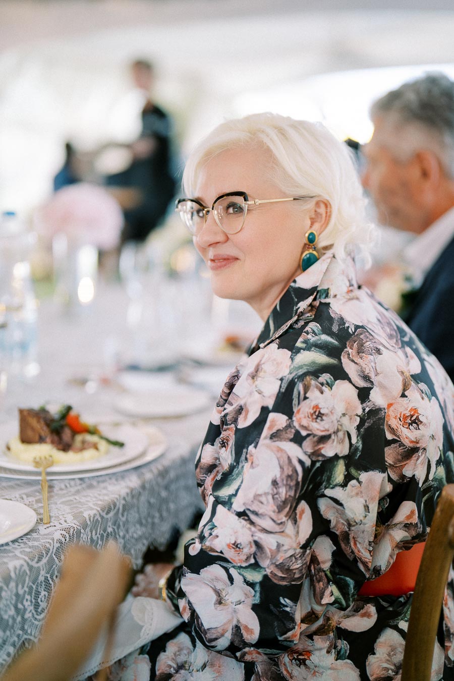 A person with short white hair and glasses, wearing a floral patterned outfit, is sitting at a table with a lace tablecloth and elegantly set dinner plate. The background is softly blurred, suggesting a formal dining setting or event.