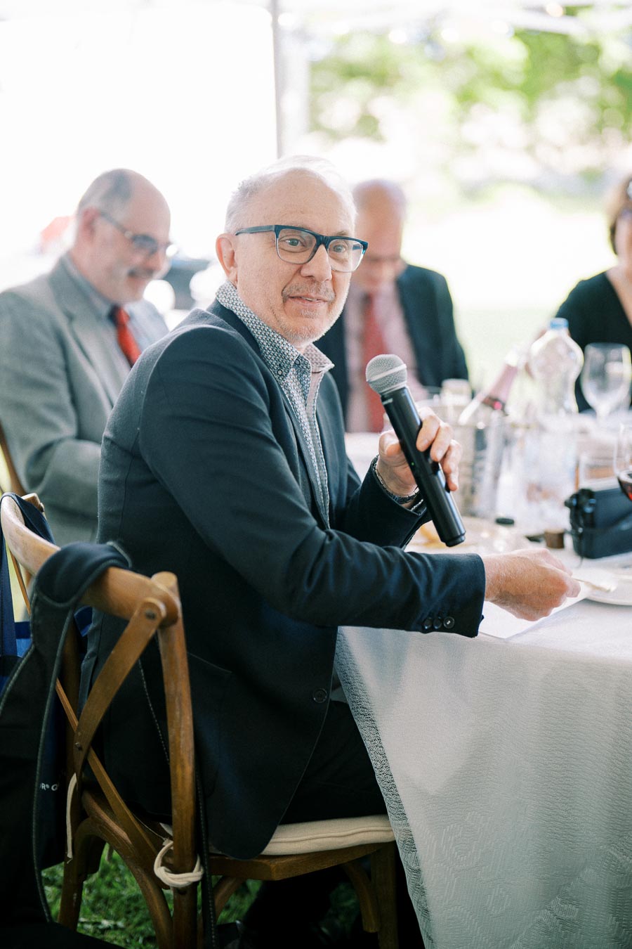 A man in a suit holding a microphone while seated at a table during an outdoor event, engaged in conversation.