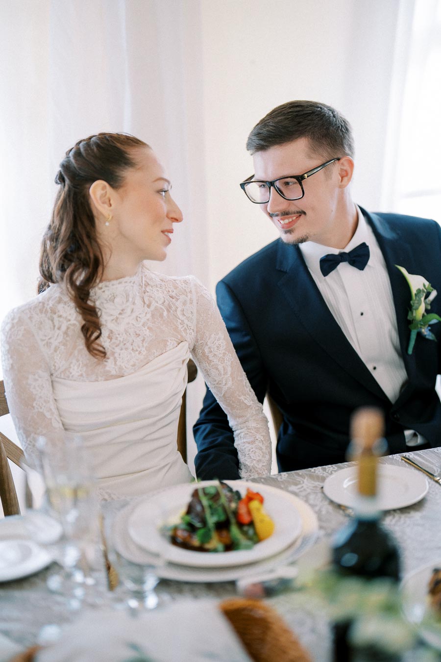 Bride and groom seated at a wedding reception table, smiling at each other. The bride wears a white lace dress, and the groom is in a dark suit with a bow tie. A plate of food and a wine bottle are on the table in front of them.
