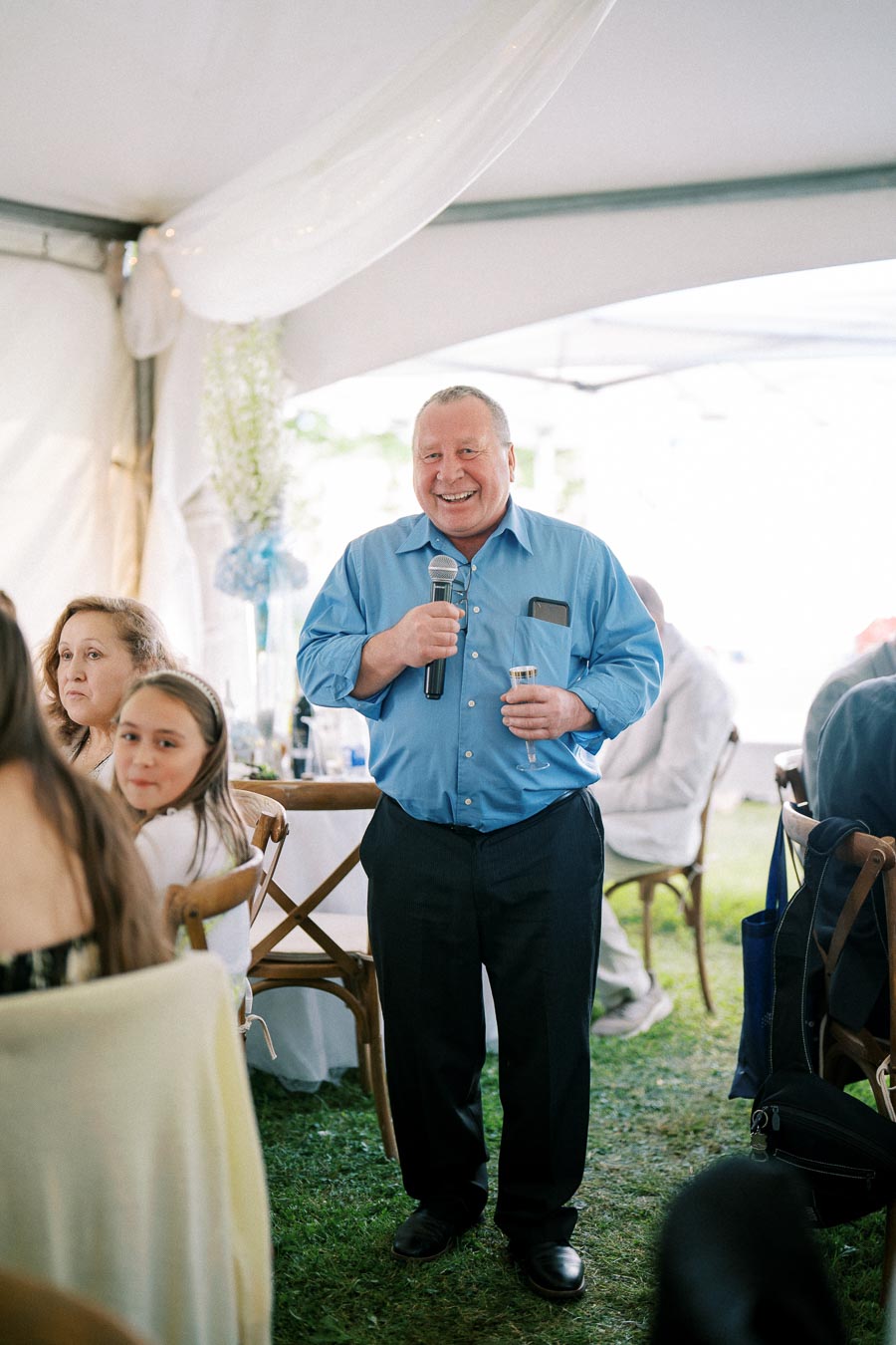 A man in a blue shirt holds a microphone and a glass, smiling while giving a speech at an outdoor event under a white tent. People are seated around him, listening attentively.