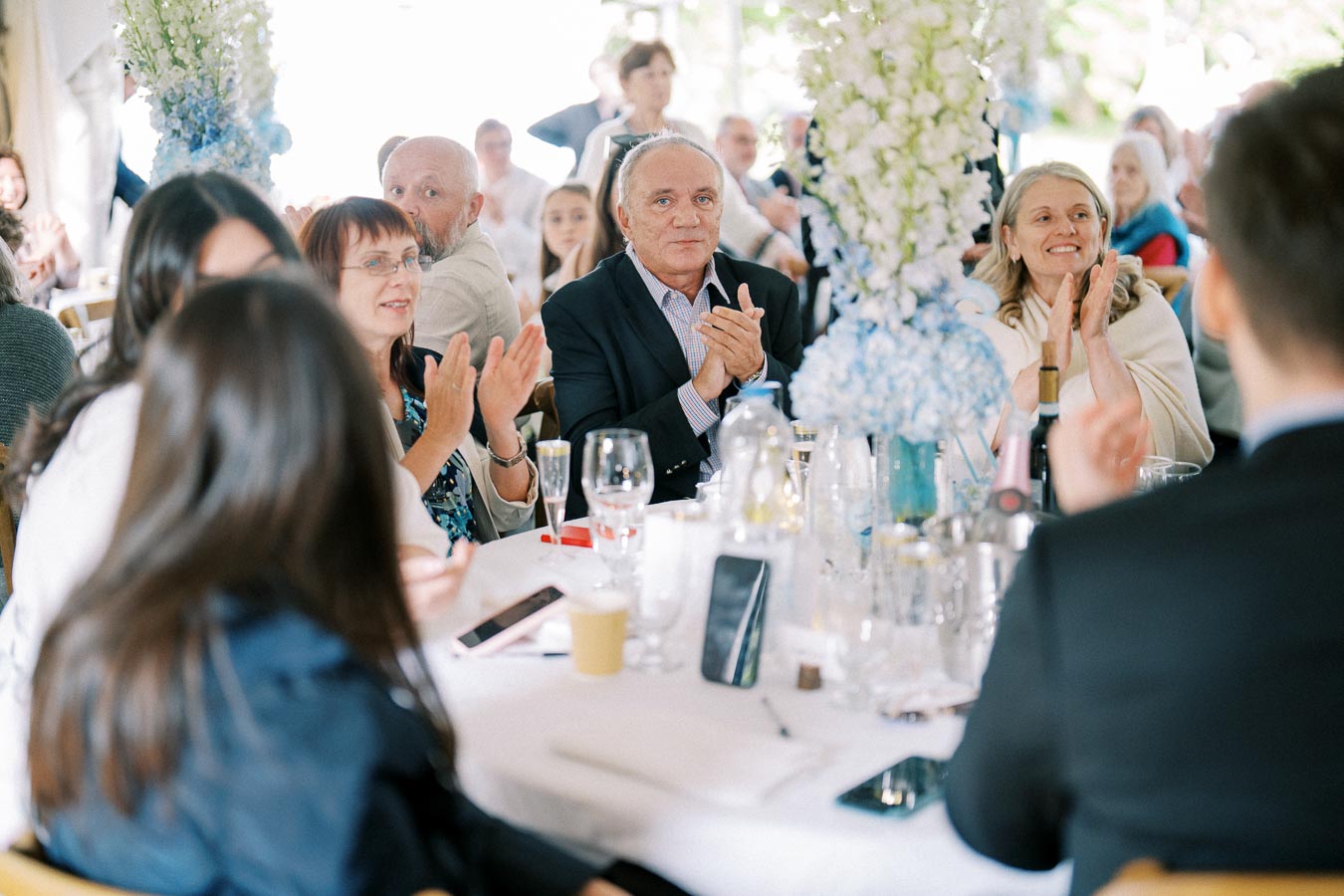 Group of people seated around a table clapping at a formal event, decorated with elegant floral arrangements and glasses on the table, creating a festive and celebratory atmosphere.