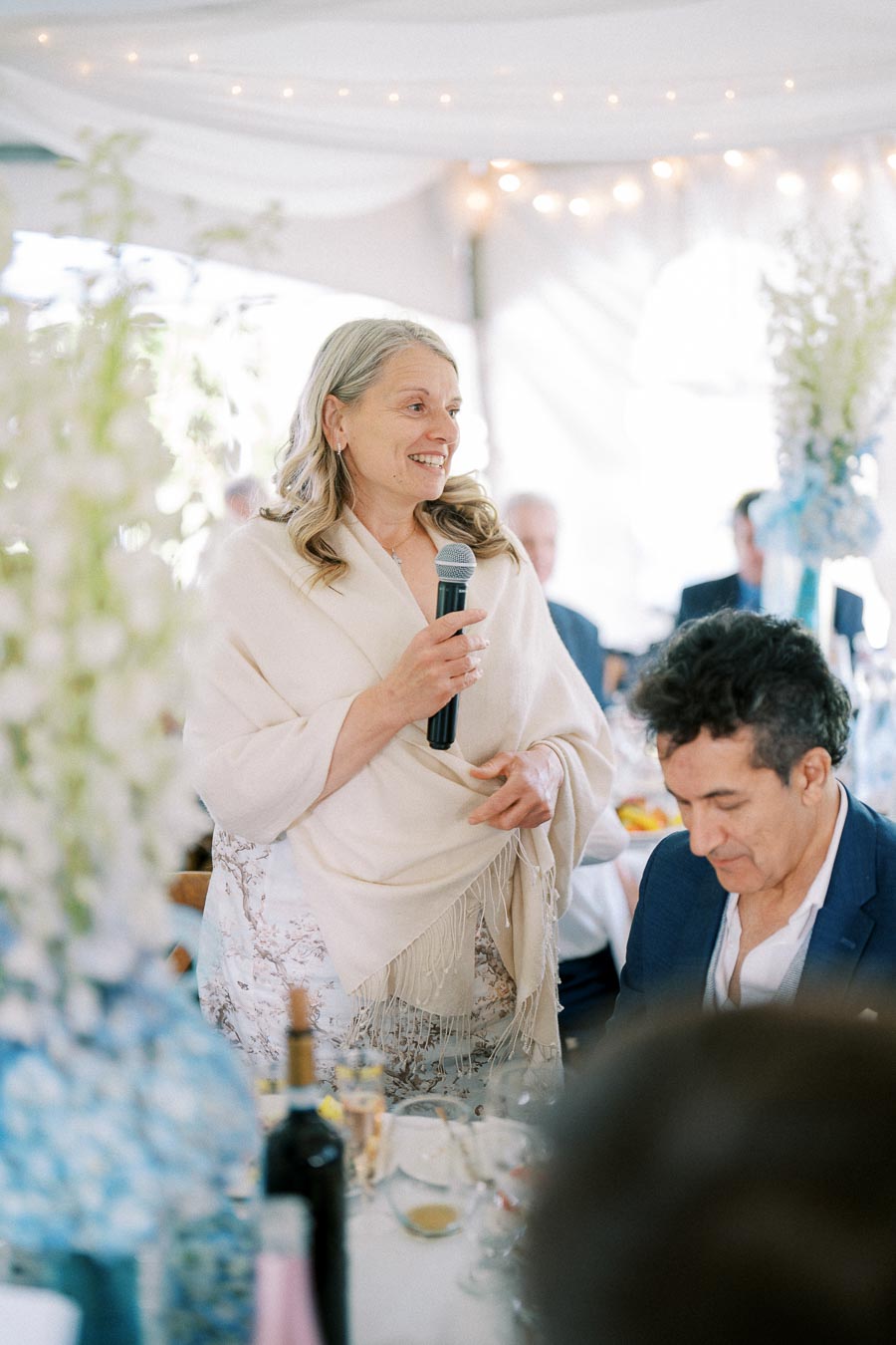 A woman in a white shawl giving a speech with a microphone at a wedding reception, surrounded by elegantly decorated tables and guests, under soft string lights.