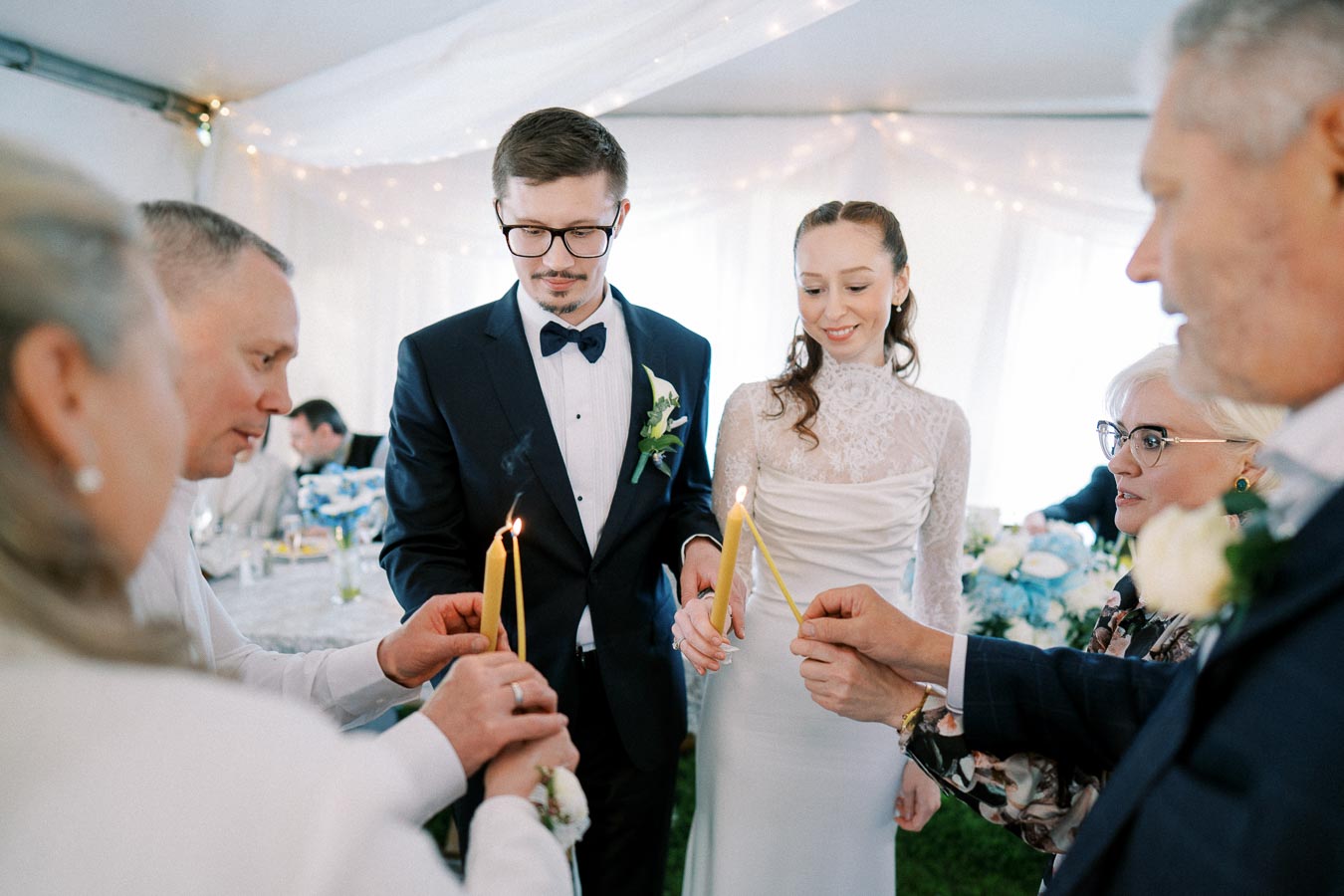 A bride and groom participate in a candle lighting ceremony with family members at a wedding, capturing a moment of unity and celebration under a beautifully decorated tent.