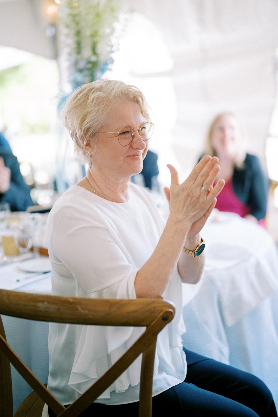 A woman with short blonde hair and glasses clapping during an event, seated at a table with a white tablecloth and guests in the background, celebrating a special occasion.