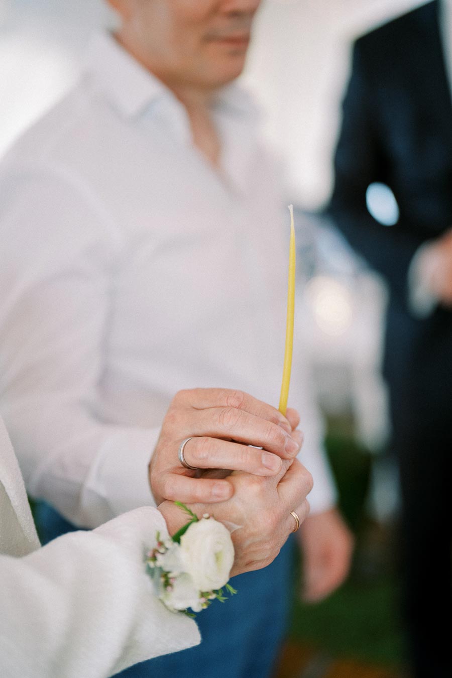 A person in a white shirt holding a thin yellow candle during a ceremony, with another hand gently holding theirs, adorned with a white floral corsage.
