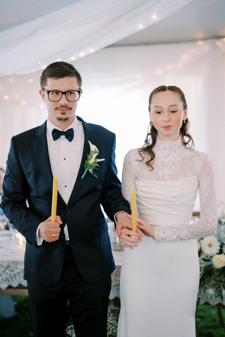 A bride and groom holding candles in a wedding ceremony, dressed in a white lace gown and a black suit with a bow tie. The setting is elegantly decorated with soft lights and floral arrangements.
