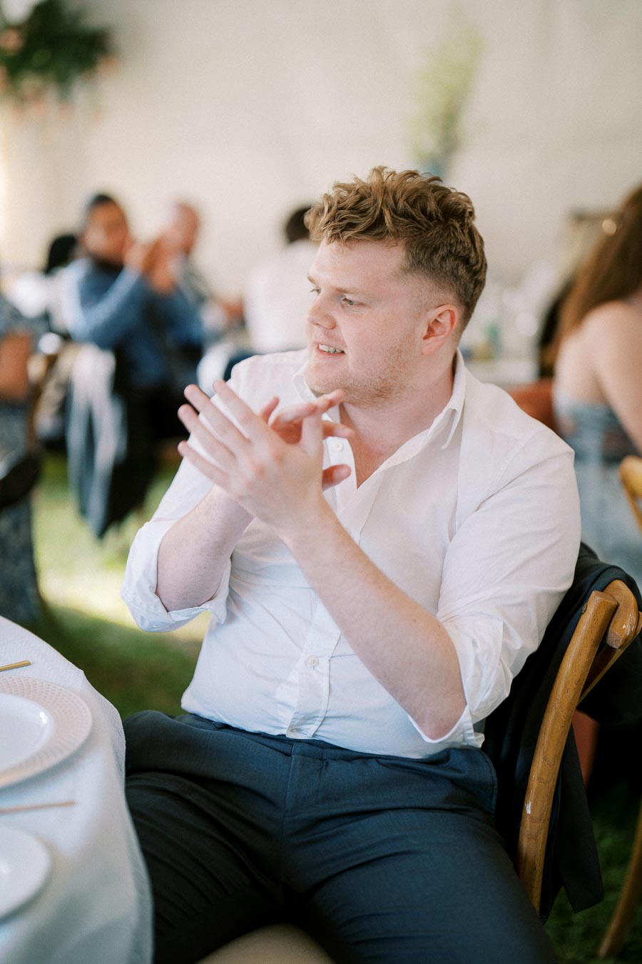 Person in a white shirt clapping at an outdoor event, seated at a table with plates, surrounded by a blurred background of people and greenery.