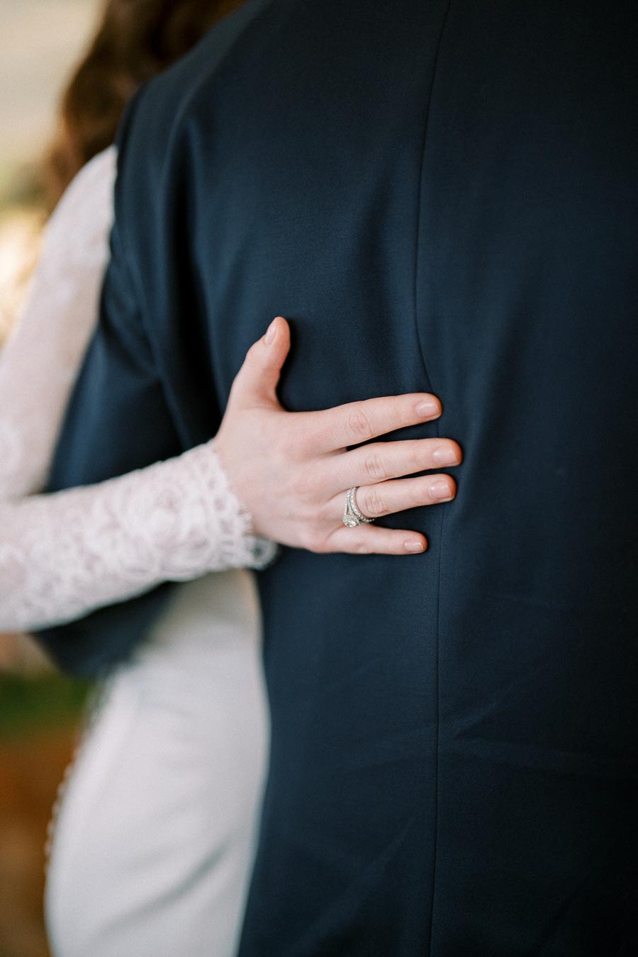 Close-up of a bride's hand with a diamond wedding ring gently embracing the groom in a dark suit, highlighting romance and elegance at a wedding ceremony.