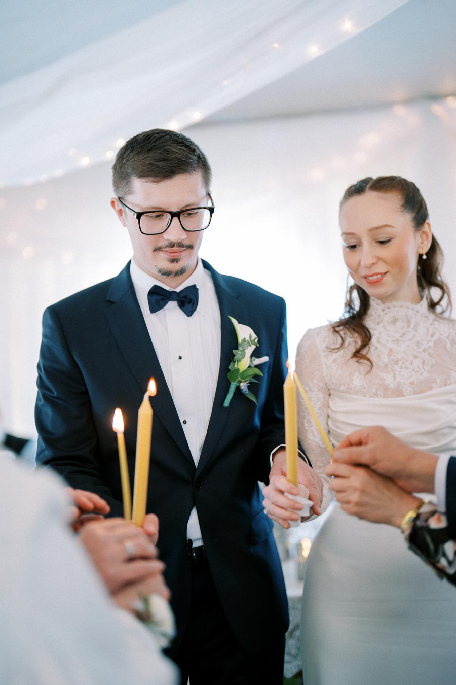 A bride and groom participate in a traditional candle lighting ceremony at their wedding, surrounded by guests in formal attire.