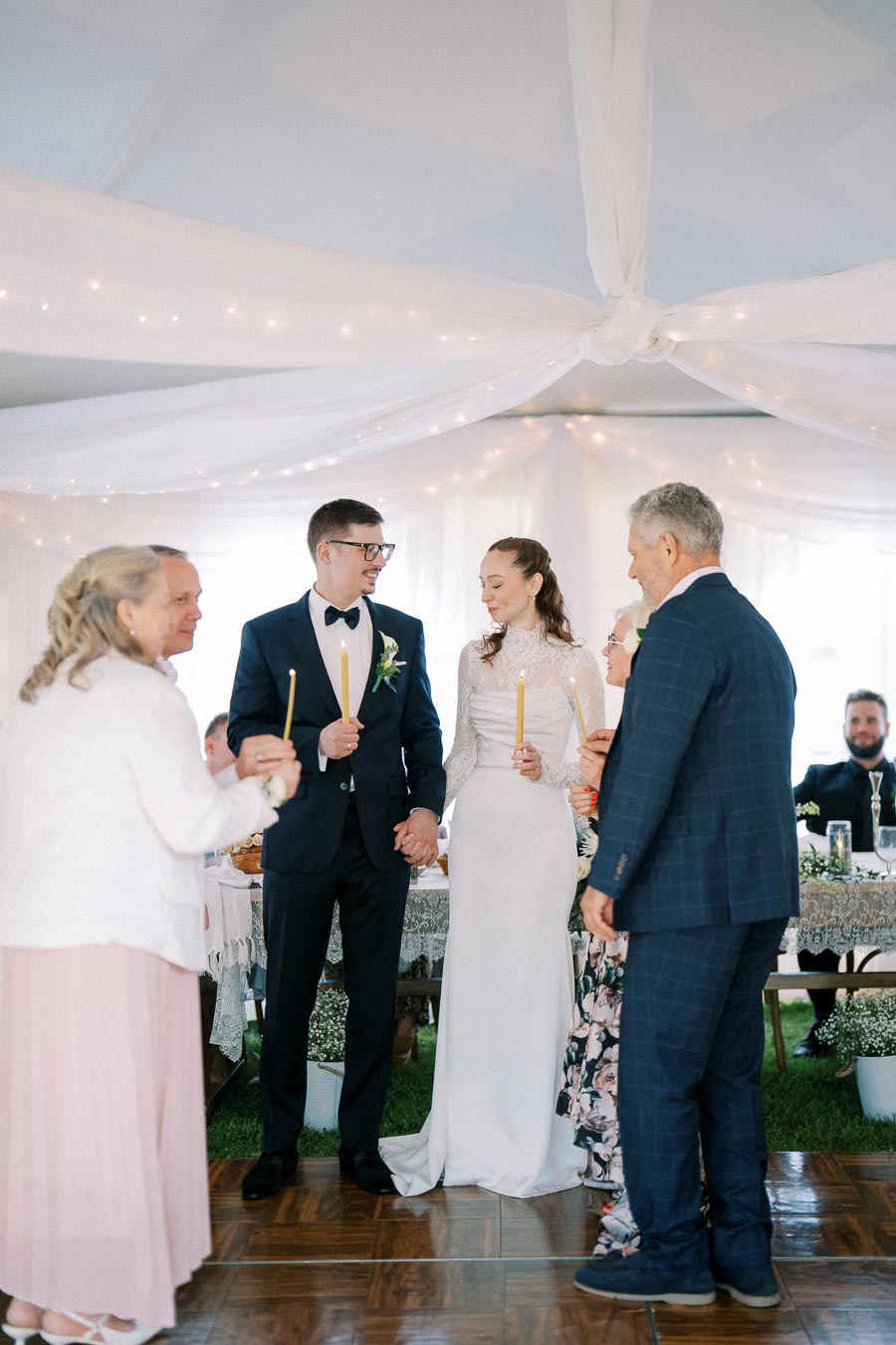A wedding ceremony under a white tent with a bride and groom holding candles, surrounded by family members, celebrating their special day in elegant attire.