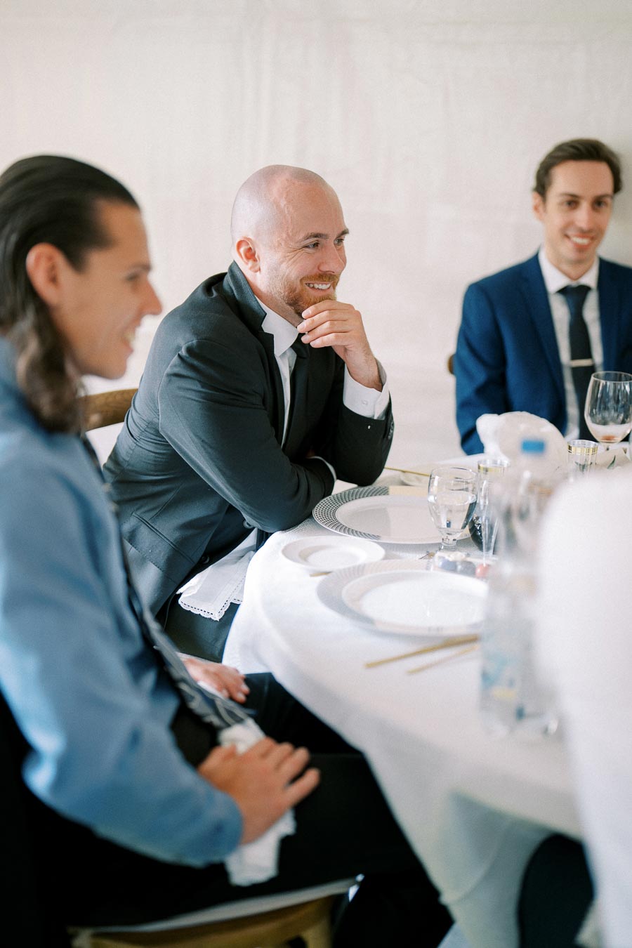 Three men in suits smiling and sitting at a round table set for a formal event.